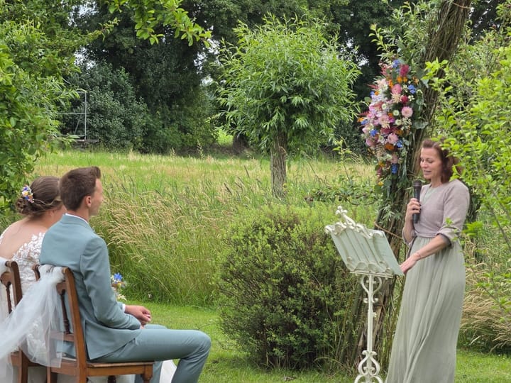 Ein Brautpaar sitzt auf der Wiese, Jasmina van Thiel spricht neben dem Blumenbogen