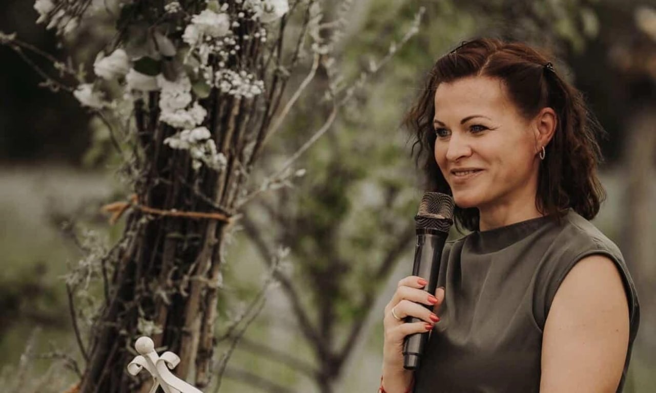 Jasmina van Thiel hält mit Mikrofon eine Traurede vor einem Blütenbogen